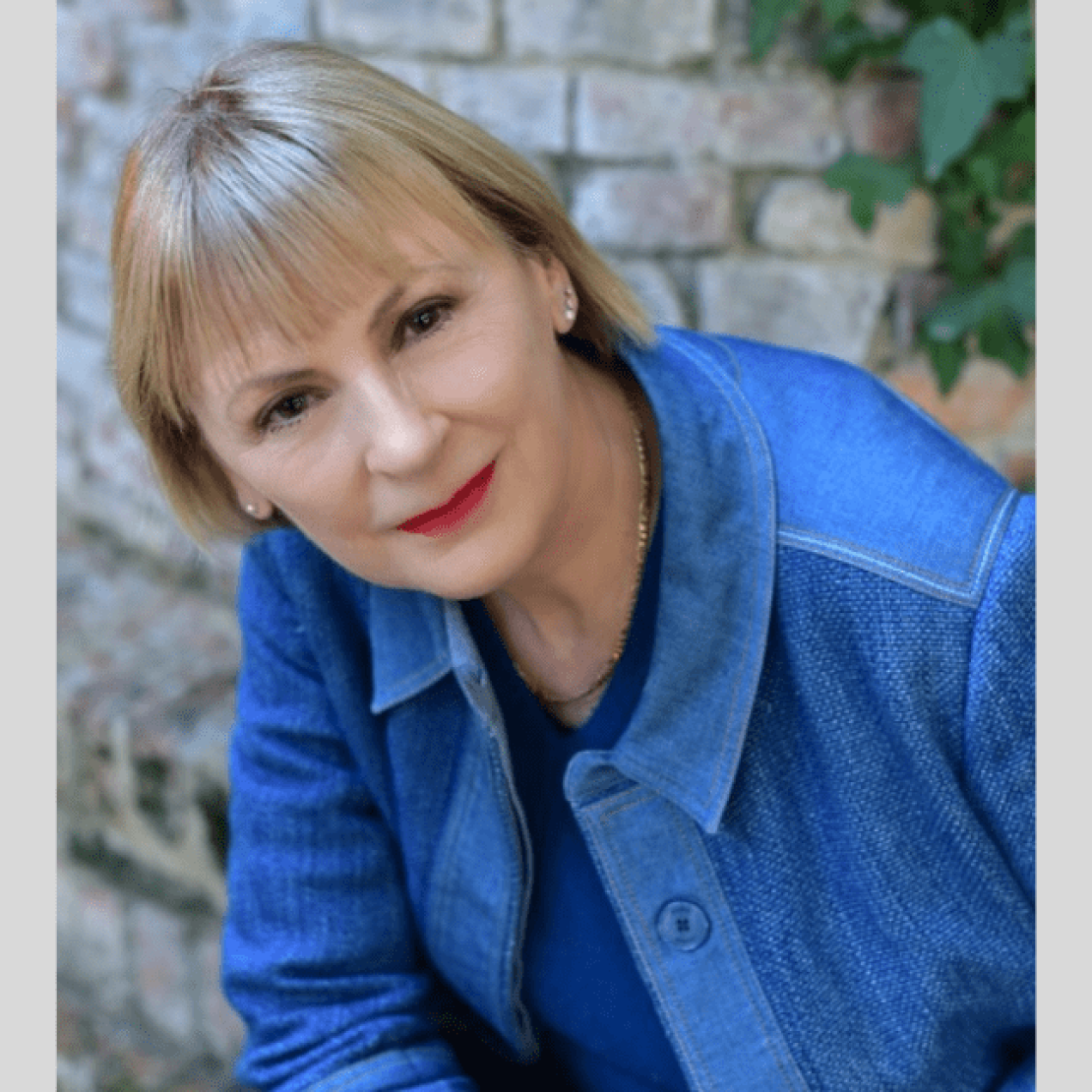 Portrait of Lindsay Nicholson, a white woman with short blonde hair wearing a blue shirt over a blue top sat with a brick wall and some foliage behind her